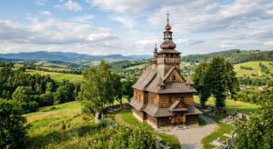 A professional editorial-style photograph of a sunlit, historic wooden church nestled in a lush, rol